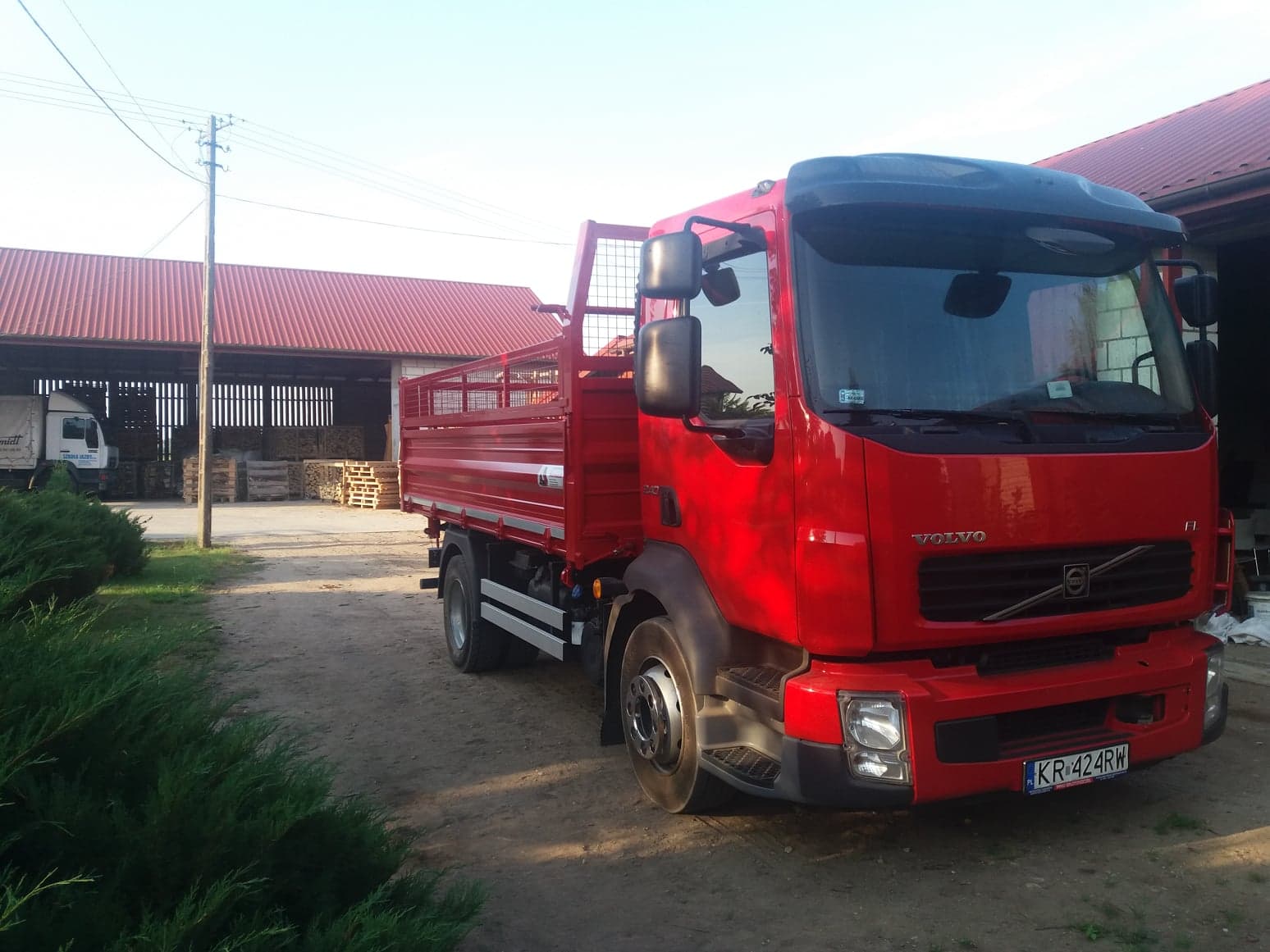 Czerwona ciężarówka Volvo z kratownicową zabudową skrzyniową na tle składu z paletami i ułożonym drewnem, widok z przodu i boku.