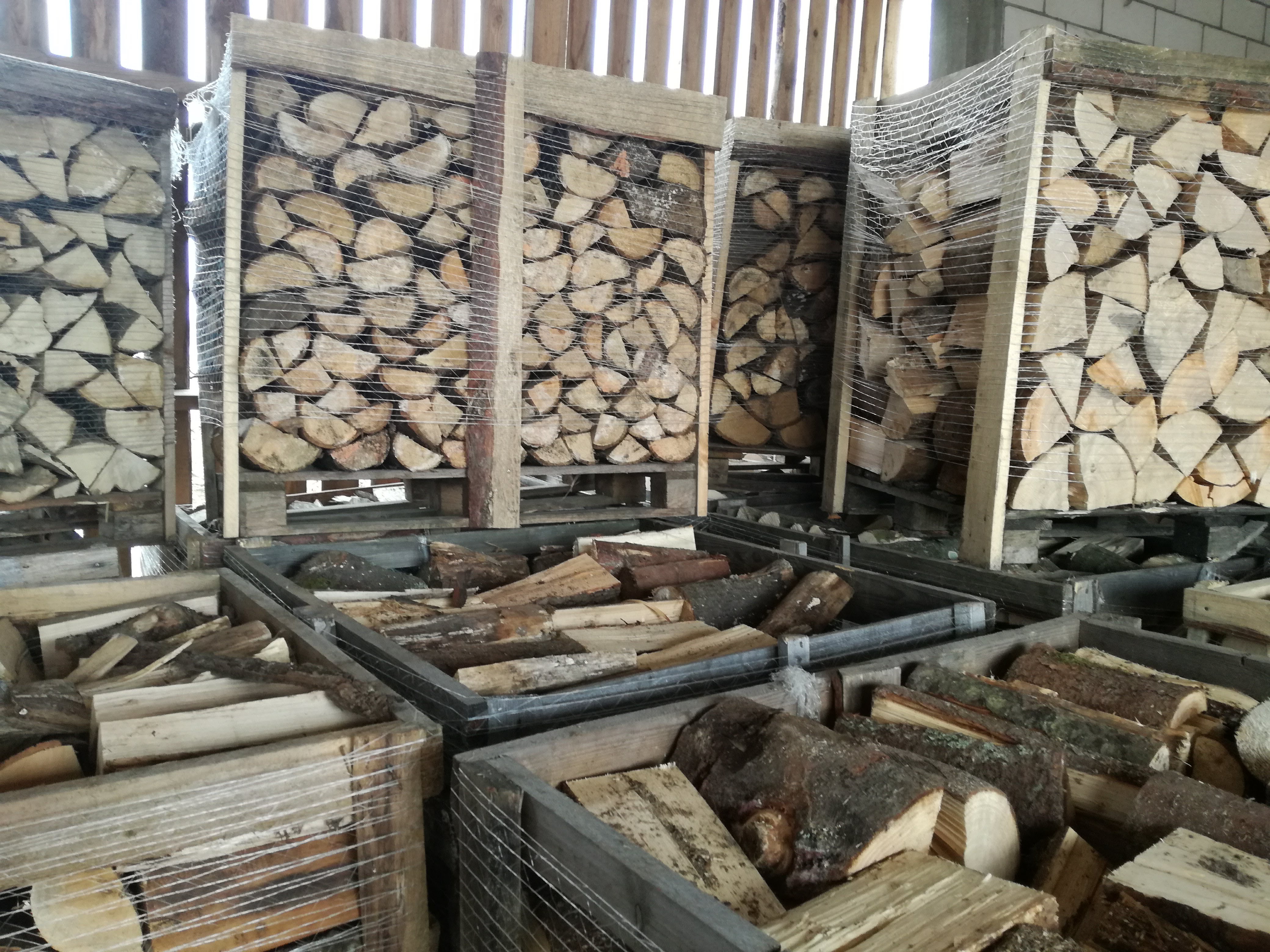 Rzędami ułożone, porąbane kawałki drewna w drewnianych skrzyniach, zabezpieczonych siatką, w magazynie.