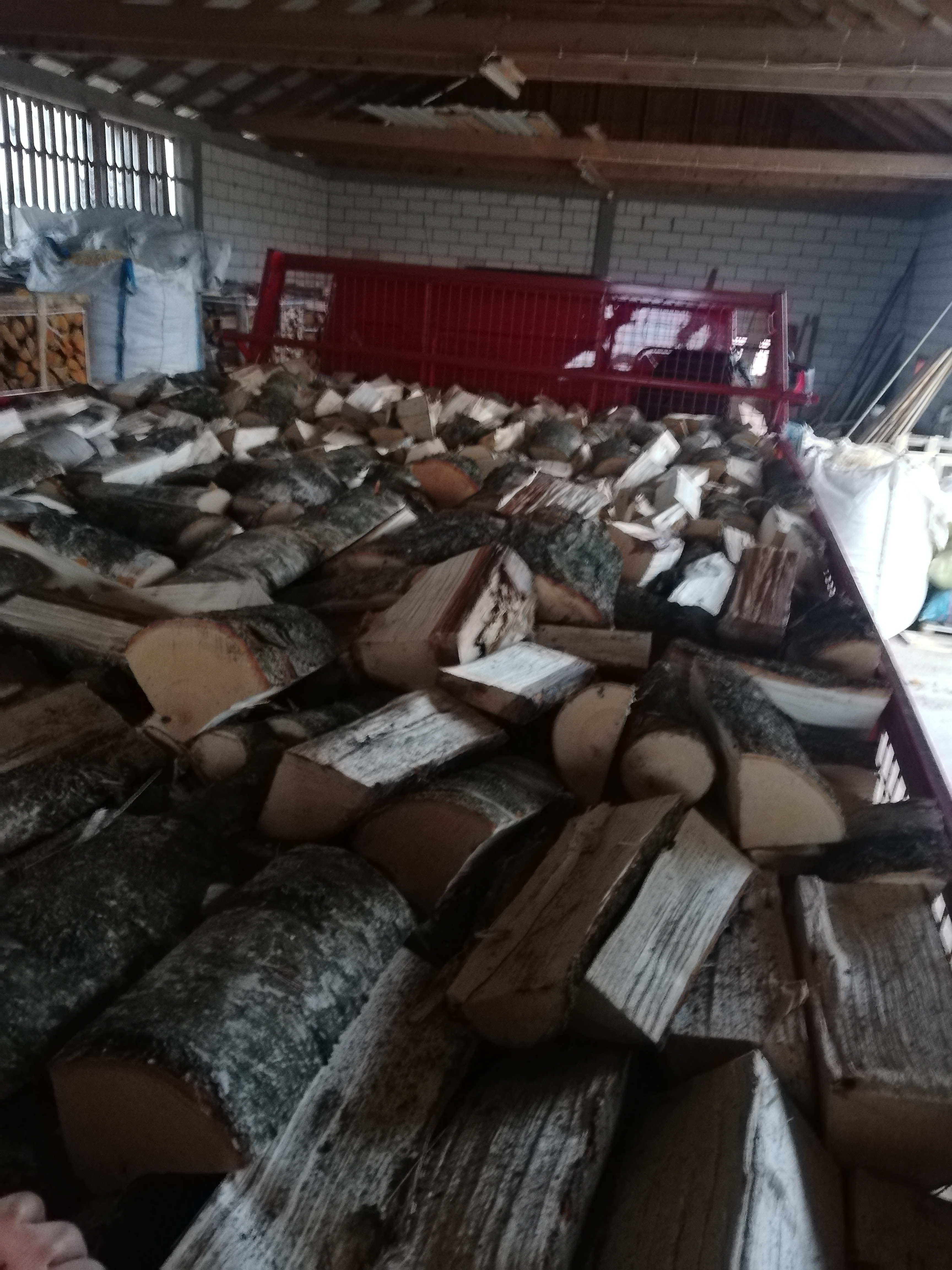 Kora brzozy na pociętych kawałkach drewna w czerwonej przyczepie, w tle budynek gospodarczy.