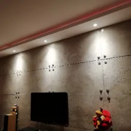 Ściana wykończona panelami imitującymi beton, połączonymi metalowymi elementami, z oświetleniem LED podsufitowym i punktowymi lampami, w tle telewizor i choinka.