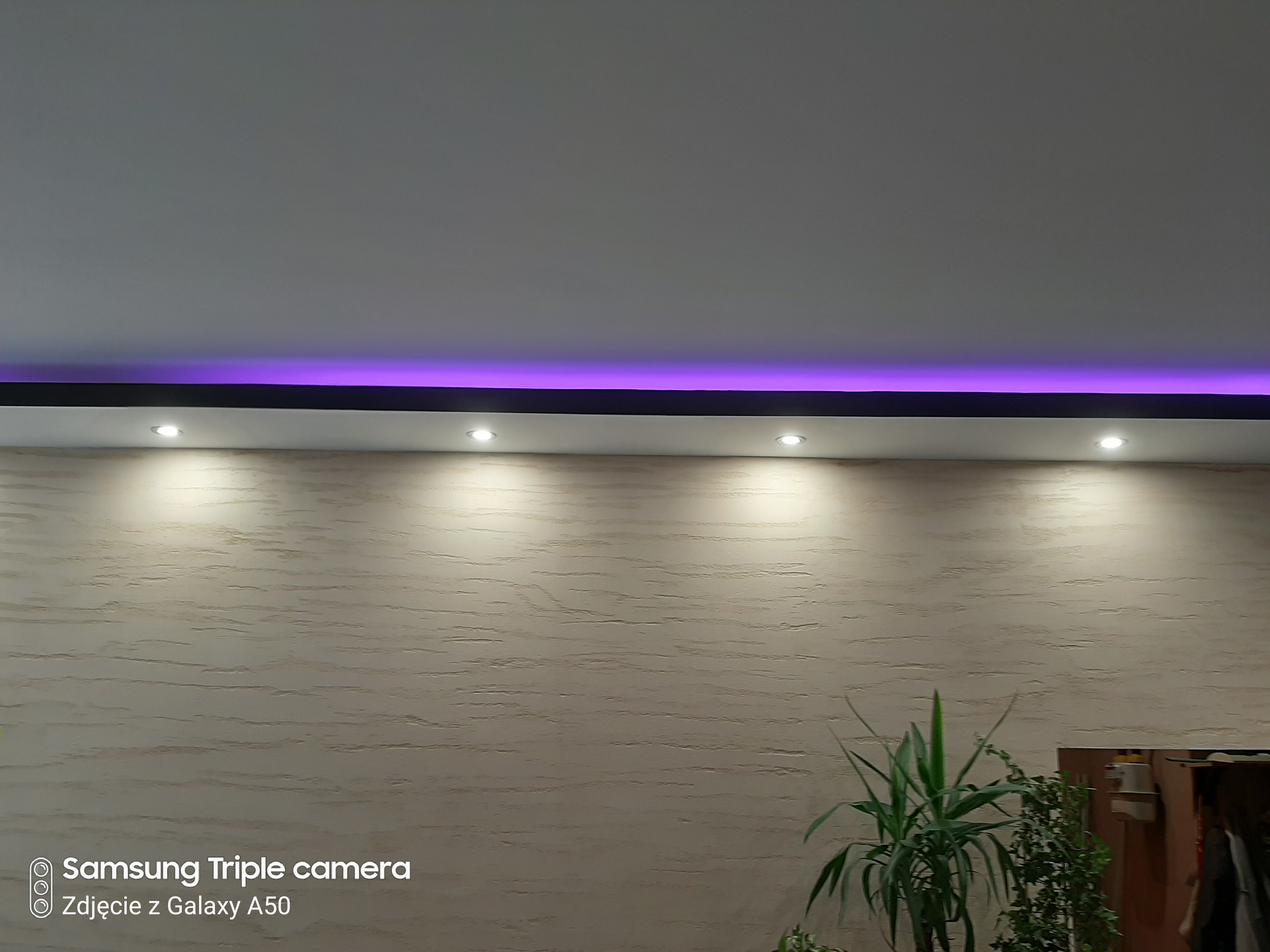 Ściana z dekoracyjną fakturą, oświetlona punktowymi lampami sufitowymi i fioletowym paskiem LED, widok z dołu.