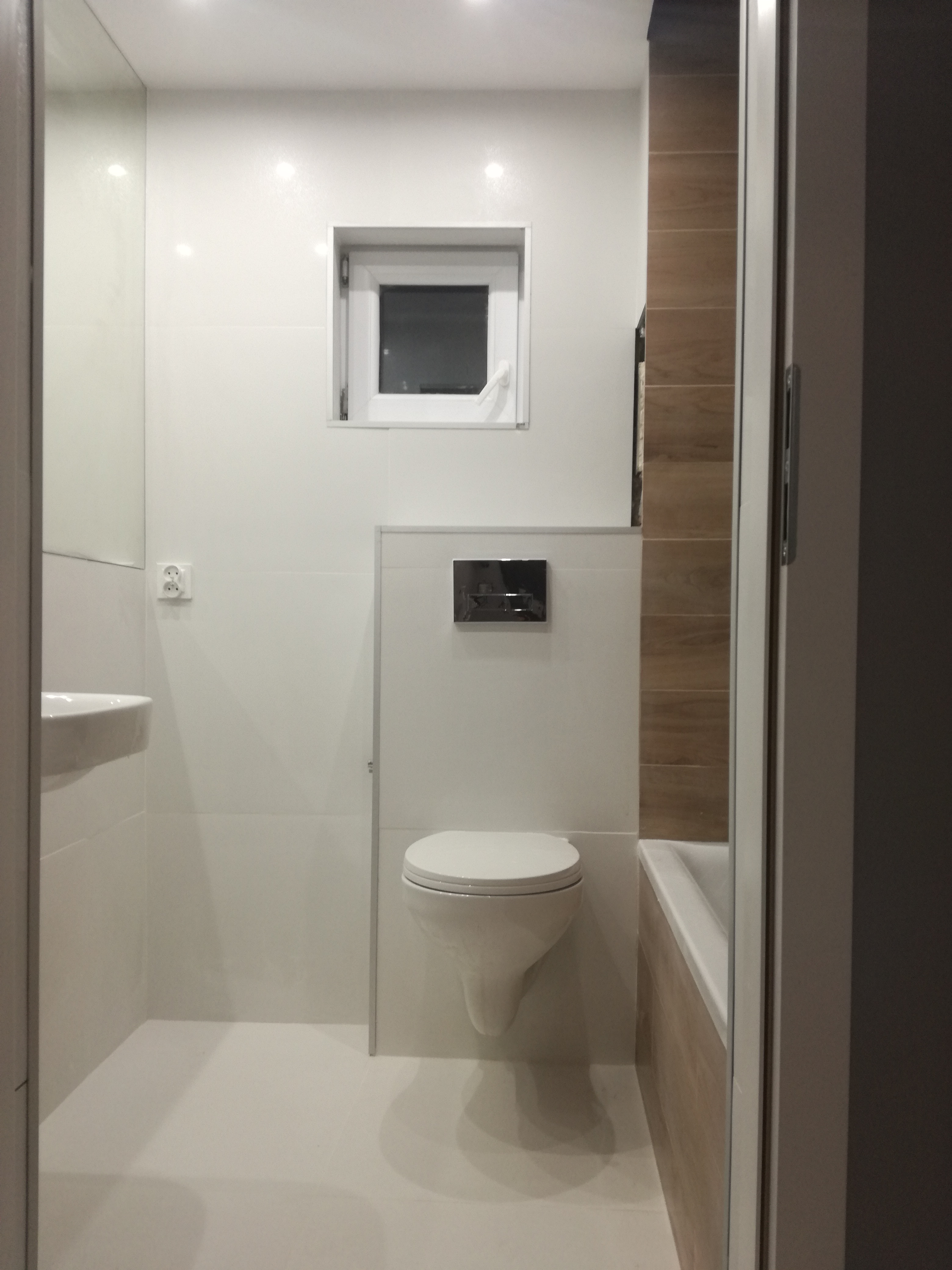 Nowoczesna, jasna łazienka z wiszącą toaletą, małym oknem i częściowo wykończoną ścianą z imitacją drewna, widziana z perspektywy drzwi.