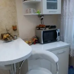 Jasna kuchnia z blatem z konglomeratu kwarcowego, białe szafki, narożna półka i hoker z chromowanymi nogami.