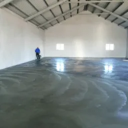 Pracownik w niebieskiej bluzie szlifuje betonową posadzkę w hali o konstrukcji stalowej i białych ścianach, widoczne refleksy światła z okien.