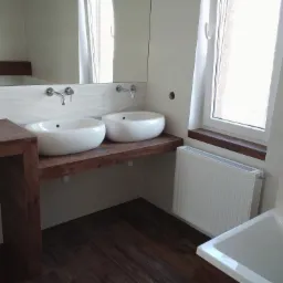 Nowoczesna łazienka z dwoma okrągłymi umywalkami nablatowymi na drewnianym blacie, białe płytki z falistym wzorem na ścianie, drewnopodobna podłoga i biały grzejnik pod oknem.