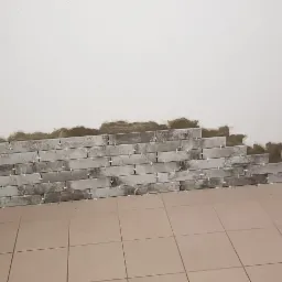 Ściana w trakcie układania szarych, cegiełkowych płytek, widoczne krzyżyki dystansowe, na podłodze beżowe płytki podłogowe.