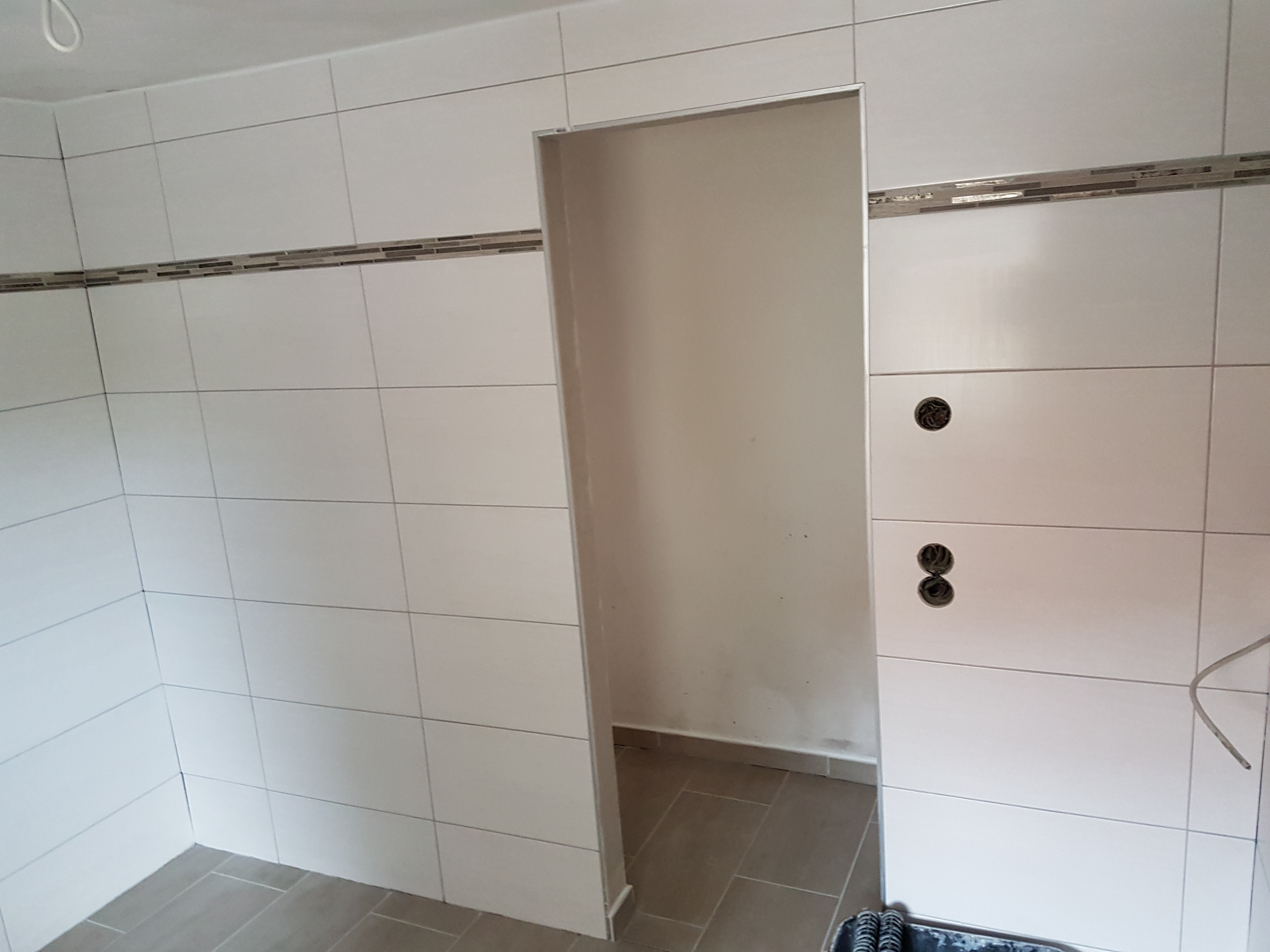 Białe, prostokątne płytki ścienne z dekoracyjnym pasem mozaiki, widoczne otwory instalacyjne i fragment podłogi z szarych płytek w wykończonej łazience z otwartym wejściem do kabiny prysznicowej.