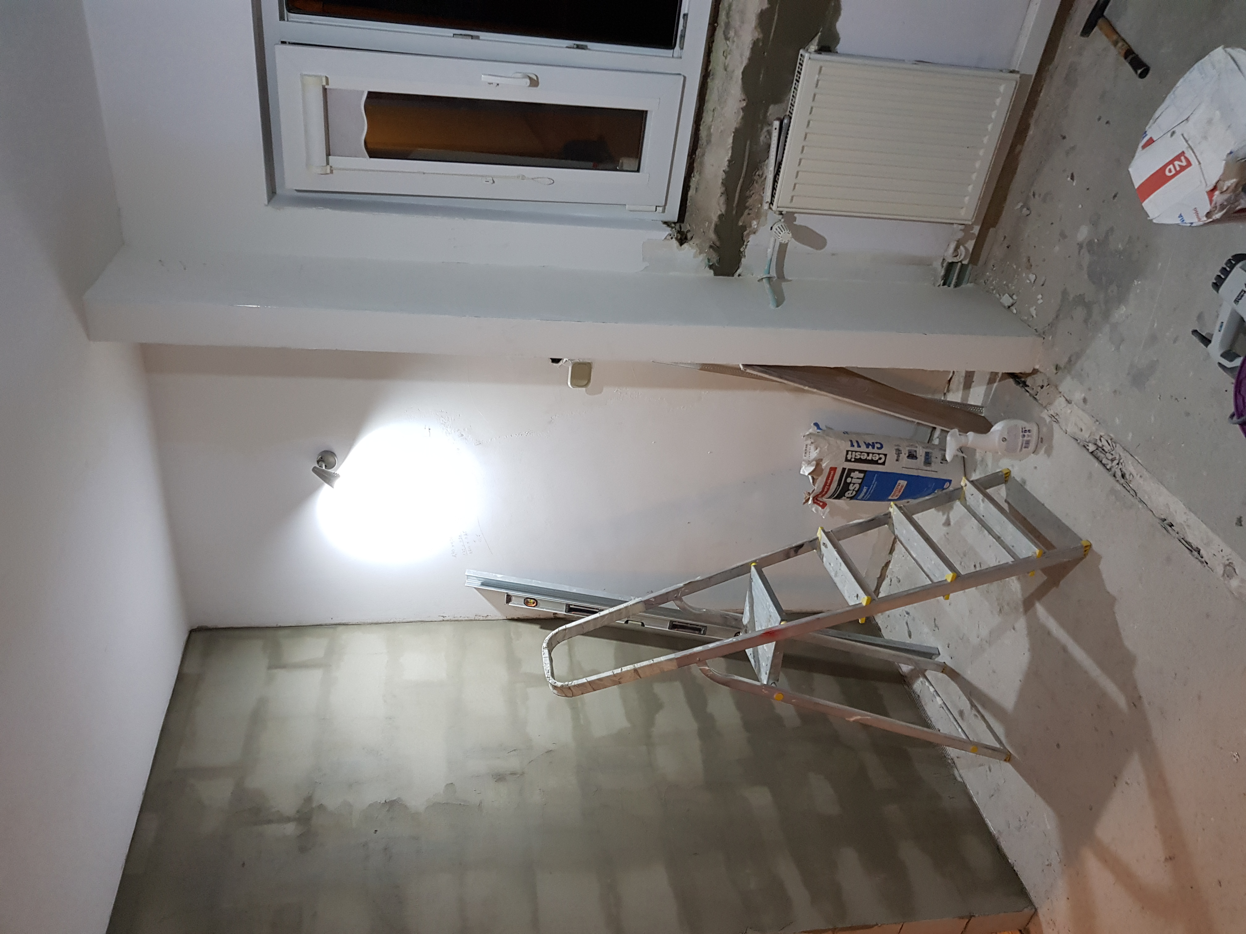 Pokój w trakcie przygotowania do malowania: surowa ściana, drabina aluminiowa, worek z zaprawą Ceresit CM 11, widoczne narzędzia i grzejnik.
