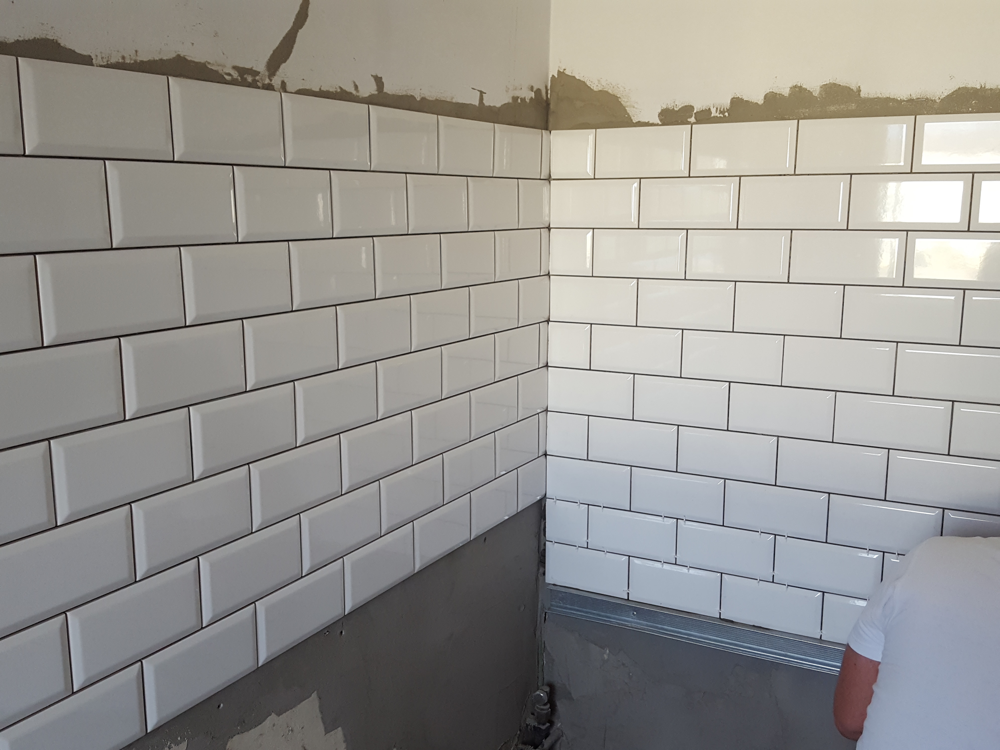 Białe, błyszczące płytki typu metro z czarną fugą, ułożone na ścianie w rogu pomieszczenia, widoczne ślady zaprawy murarskiej na górze ściany, dolne rzędy płytek w trakcie montażu, plastikowe...