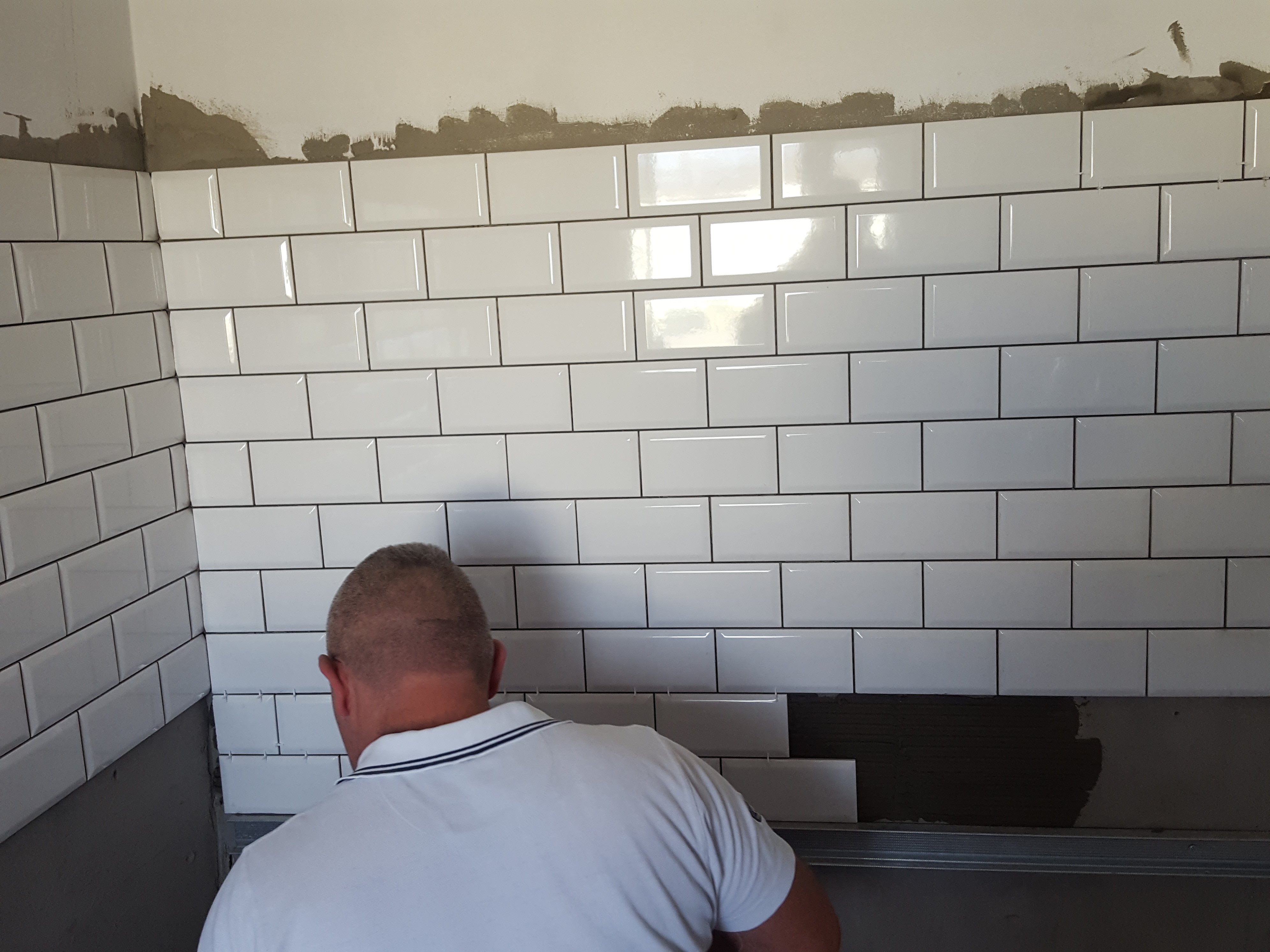 Układanie białych, prostokątnych płytek na ścianie z ciemną fugą; widoczny fragment ściany bez płytek i osoba w trakcie pracy.