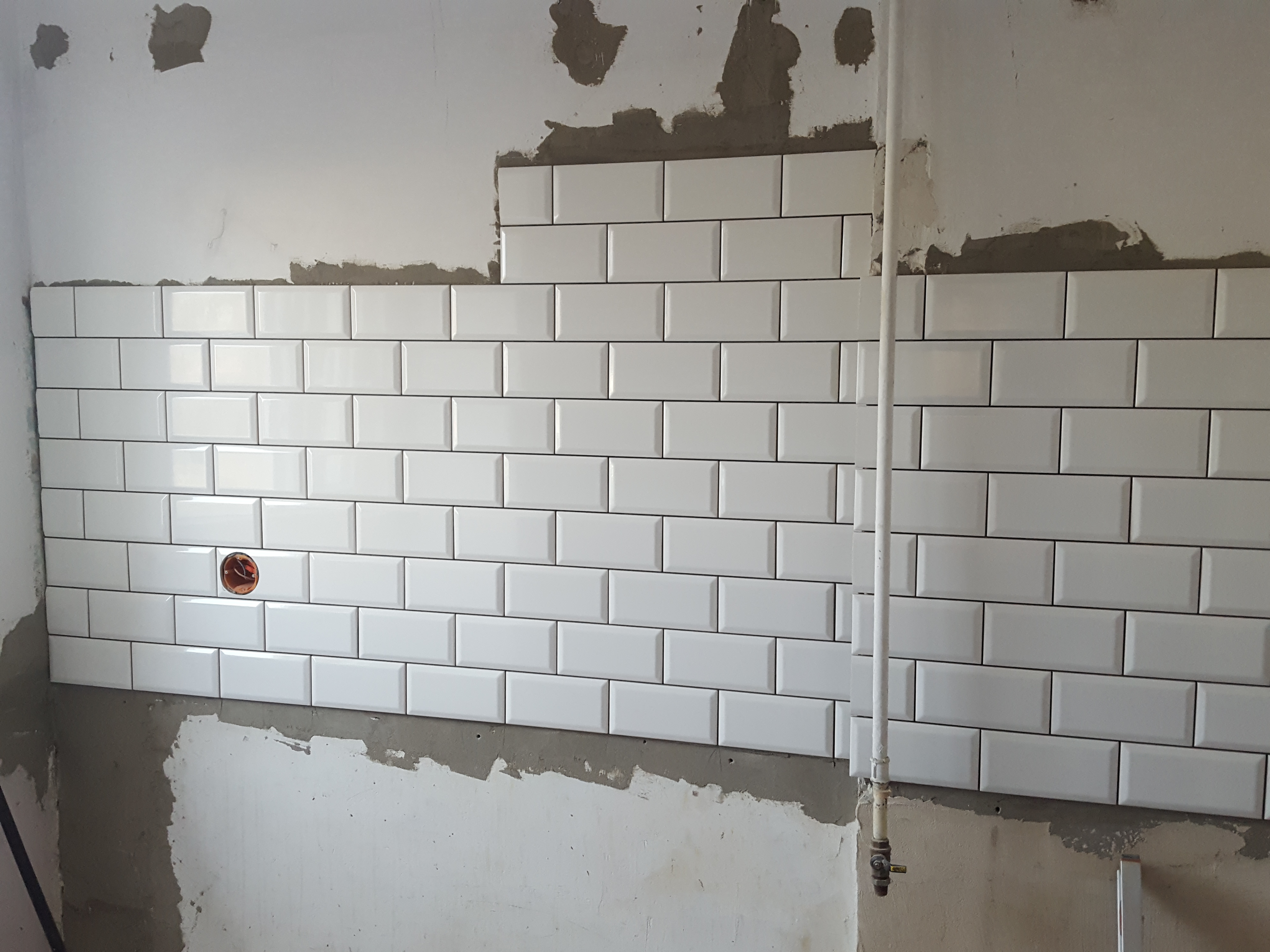 Ściana w trakcie układania białych, prostokątnych płytek typu metro, z widocznym gniazdkiem elektrycznym i fragmentami nieotynkowanej ściany powyżej oraz rurą pionową po prawej stronie.