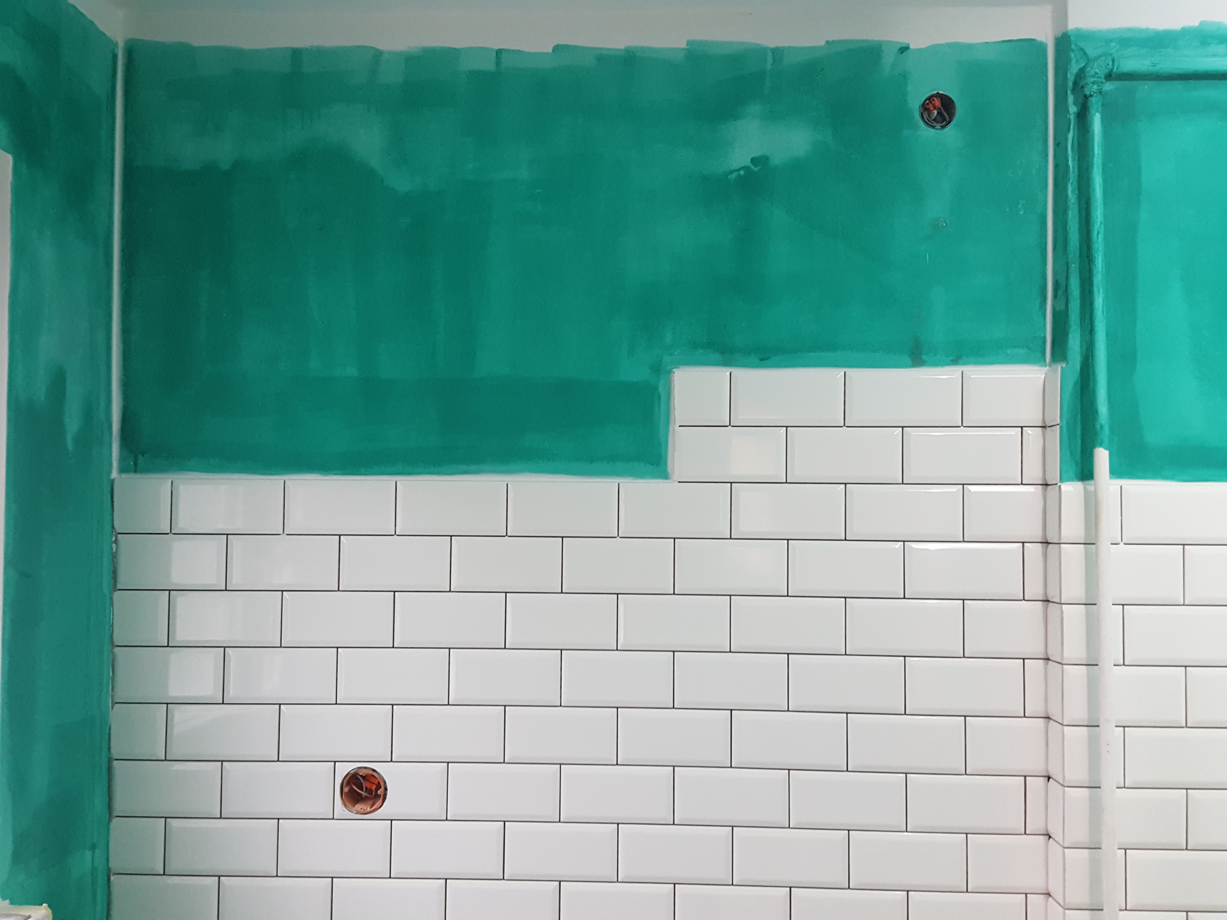 Ściana w trakcie remontu: dolna część wyłożona białymi, prostokątnymi płytkami, górna część pomalowana na zielono, widoczne otwory na gniazdka elektryczne.