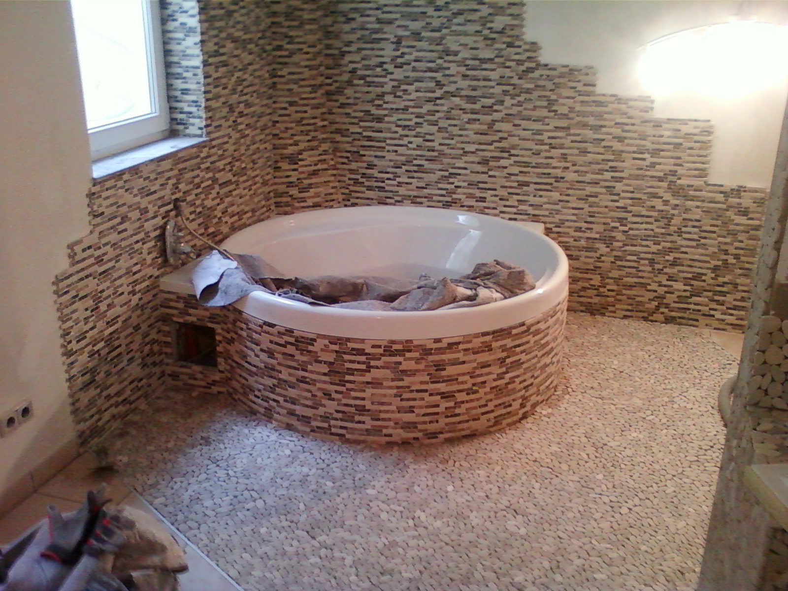 Okrągła wanna obłożona mozaiką w łazience z podłogą wyłożoną drobnymi kamieniami, ściany częściowo wykończone mozaiką, zabezpieczona folią malarską.