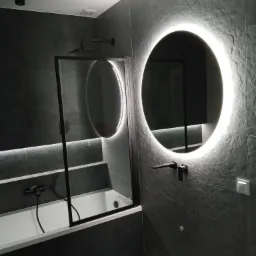 Nowoczesna łazienka z czarnymi płytkami imitującymi kamień, podświetlanym okrągłym lustrem, czarną armaturą i wanną z czarną ramą parawanu.