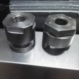 Dwa metalowe, cylindryczne elementy z gwintem wewnętrznym i sześciokątną podstawą, leżące na metalowej powierzchni, z widocznymi śladami obróbki.