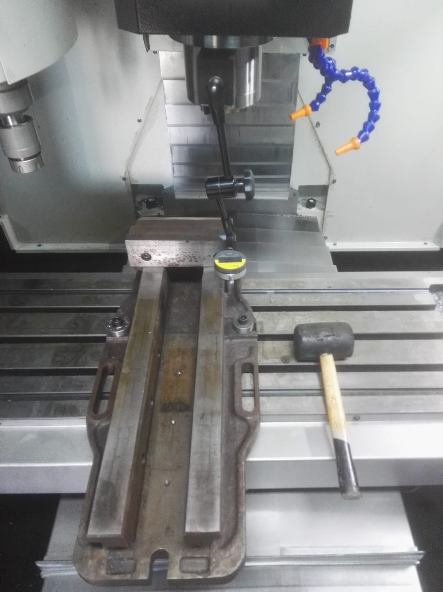 Ustawianie uchwytu obrabianego elementu w centrum roboczym frezarki CNC z użyciem czujnika zegarowego; w tle widoczne dysze chłodziwa i młotek.