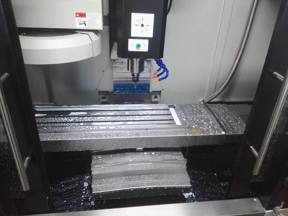 Wnętrze maszyny CNC podczas obróbki metalu, widoczny frez, chłodziwo i obrabiany element z aluminium.