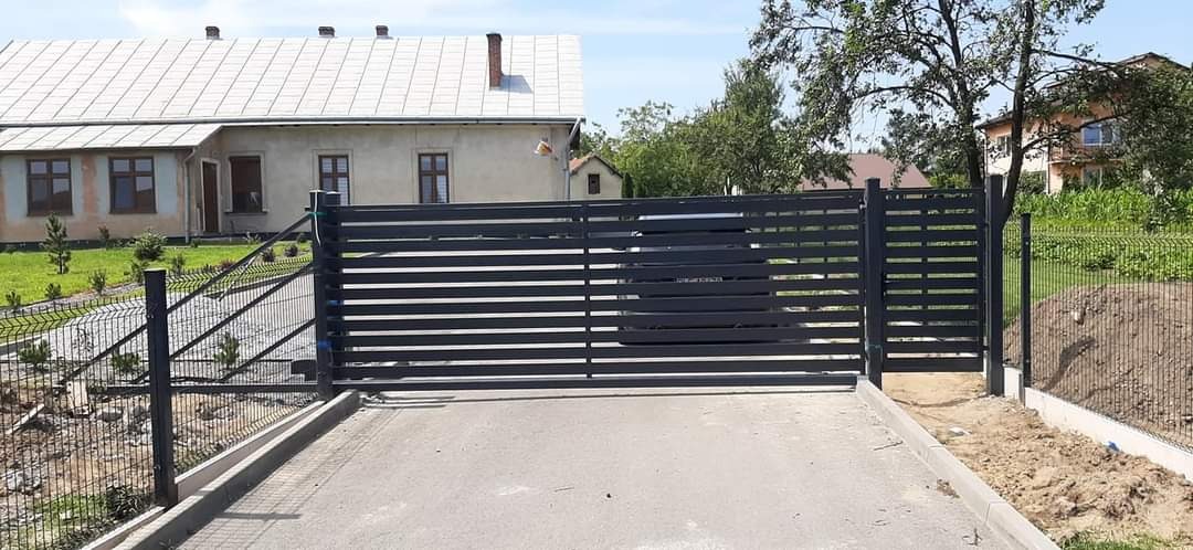 Nowoczesna, przesuwna brama posesyjna z poziomych profili, pomalowana na ciemny kolor, z widoczną siatką ogrodzeniową i budynkiem mieszkalnym w tle.