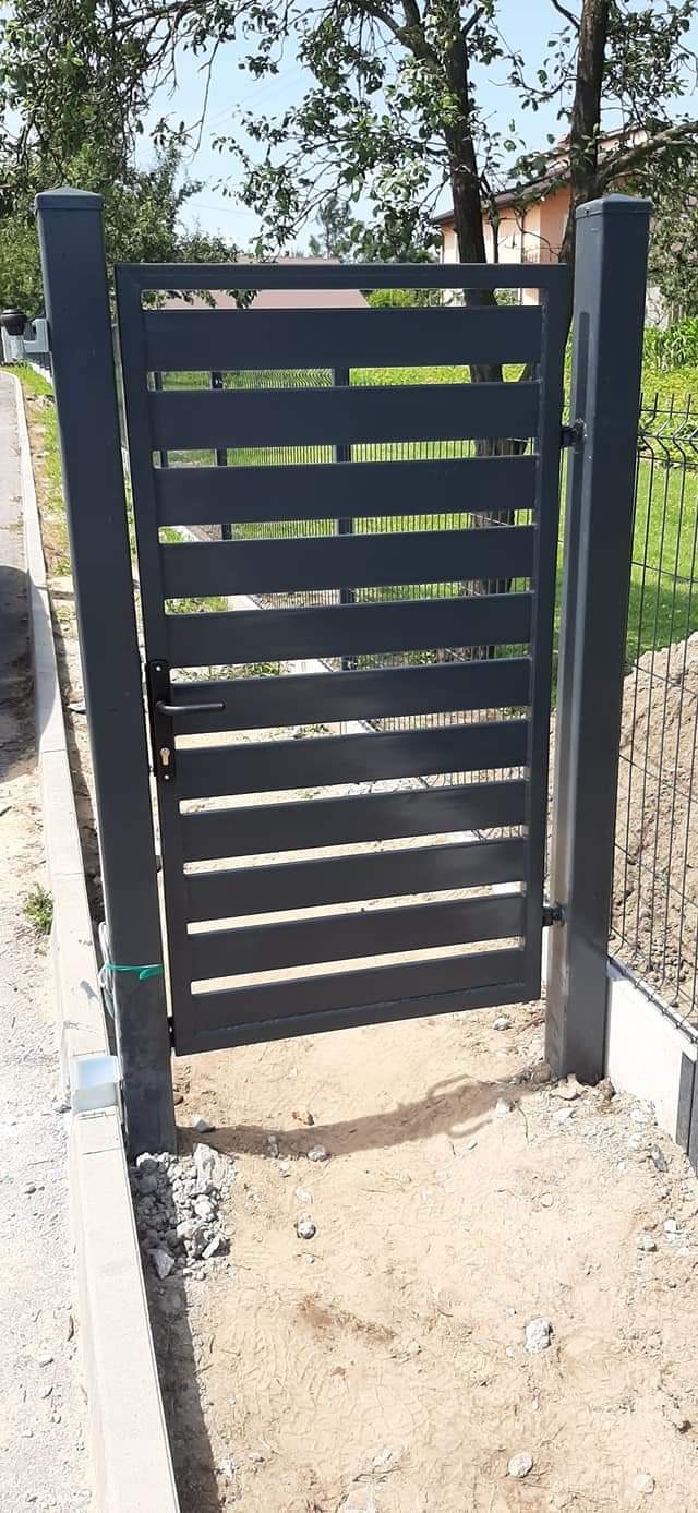 Nowoczesna, antracytowa furtka z poziomych profili stalowych, zamontowana na słupkach, widok z zewnątrz posesji, teren wokół furtki jeszcze niezagospodarowany.