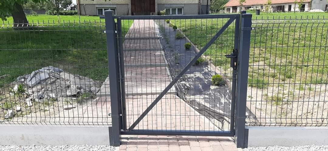 Nowoczesna, metalowa furtka panelowa w kolorze grafitowym, zamykana na klamkę, prowadząca do posesji z brukowaną ścieżką i trawnikiem w tle.