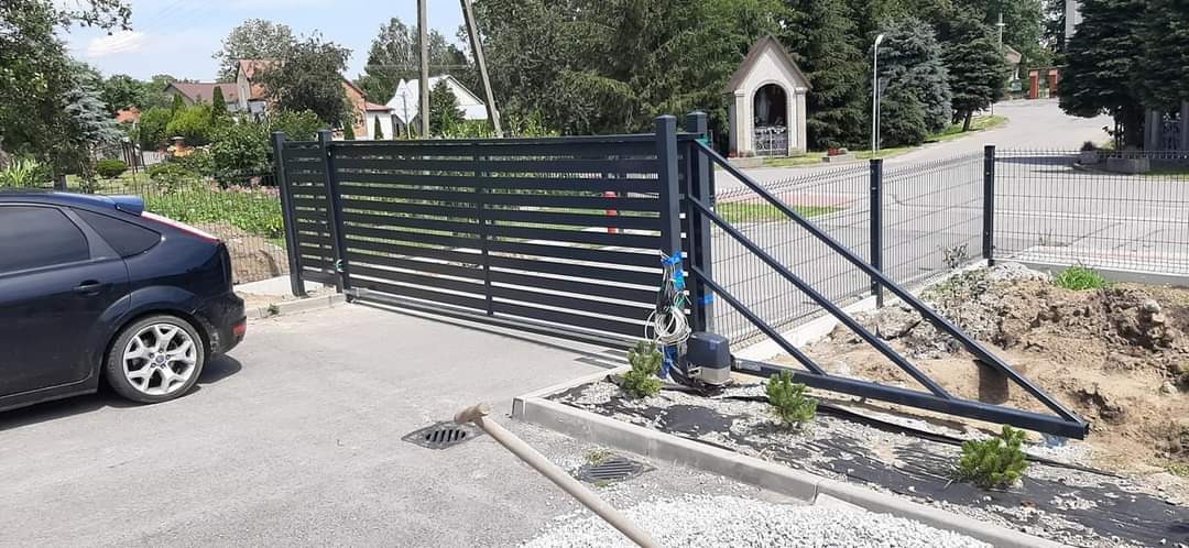Nowoczesna, pozioma brama przesuwna z automatyką, widok z zewnątrz, w tle kapliczka i zabudowa jednorodzinna.