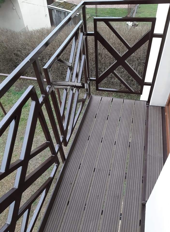 Balkon z brązową, metalową balustradą o geometrycznym wzorze i drewnianą podłogą, widok z góry, brama otwarta.