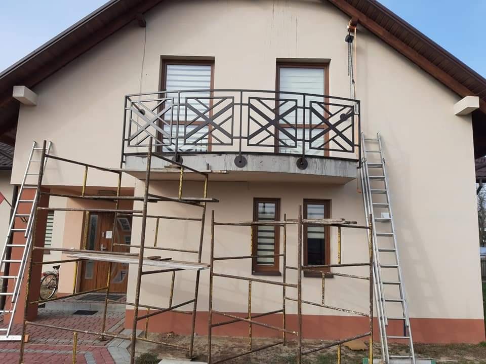 Montaż balustrady balkonowej z motywem geometrycznym na budynku jednorodzinnym, widoczne rusztowania i drabiny.