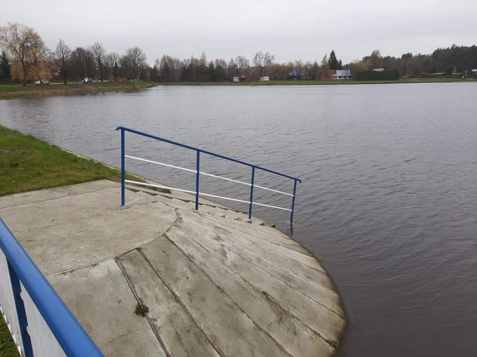 Betonowe schody z niebiesko-białą barierką prowadzące do szarego jeziora w pochmurny dzień.
