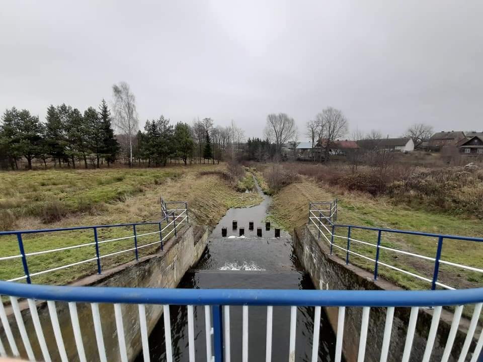 Betonowa konstrukcja z niebiesko-białą barierką nad kanałem wodnym z rzędem czarnych słupków spowalniających przepływ, widok z góry na tle krajobrazu z drzewami i zabudowaniami w pochmurny dzień.