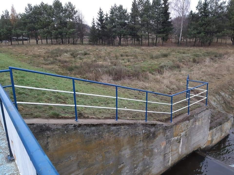 Niebiesko-biała barierka ochronna zamontowana na betonowej konstrukcji przy brzegu rzeki, widok na zalesiony teren w tle.