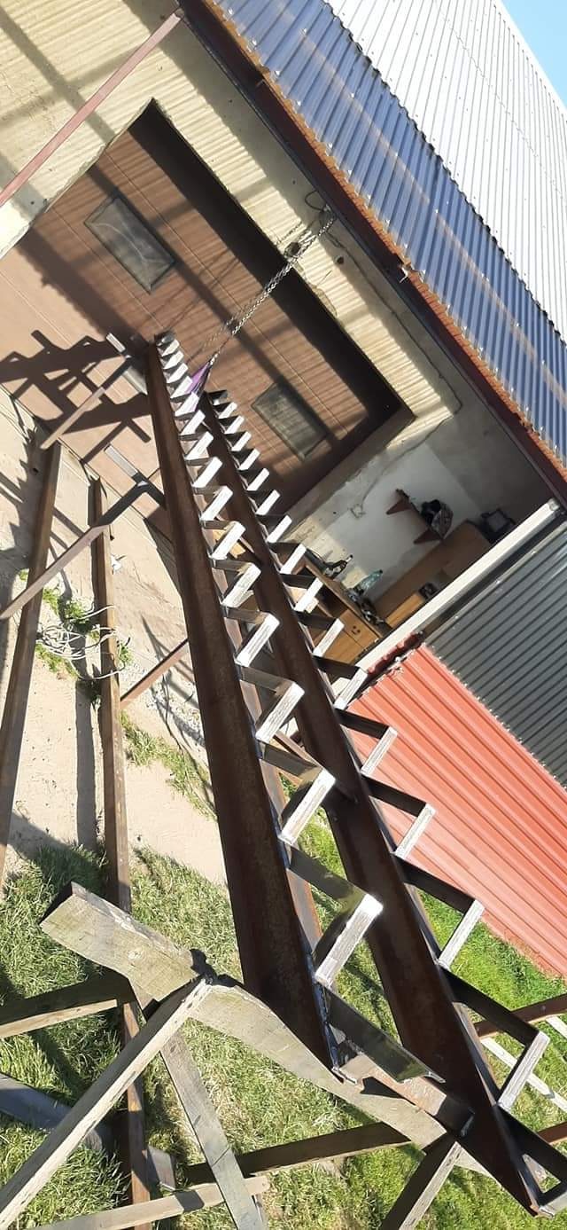 Konstrukcja metalowej drabiny z przyspawanymi szczeblami, w trakcie budowy, widok z dołu w kierunku budynku z czerwonym dachem.