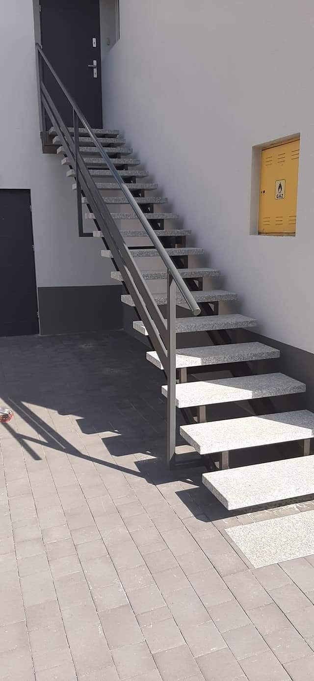 Zewnętrzne schody z szarego granitu z metalową balustradą prowadzące do drzwi wejściowych, obok żółta skrzynka gazowa.