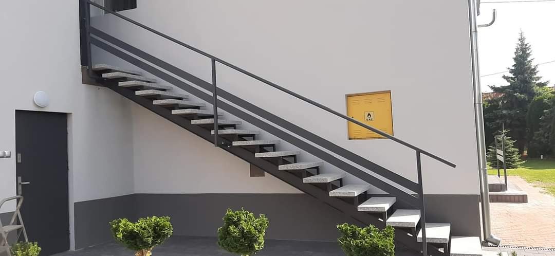 Zewnętrzne schody z szarymi, granitowymi stopniami i metalową, ciemnoszarą balustradą, prowadzące do budynku z żółtą skrzynką gazową na ścianie. U dołu widoczne małe, okrągłe krzewy.