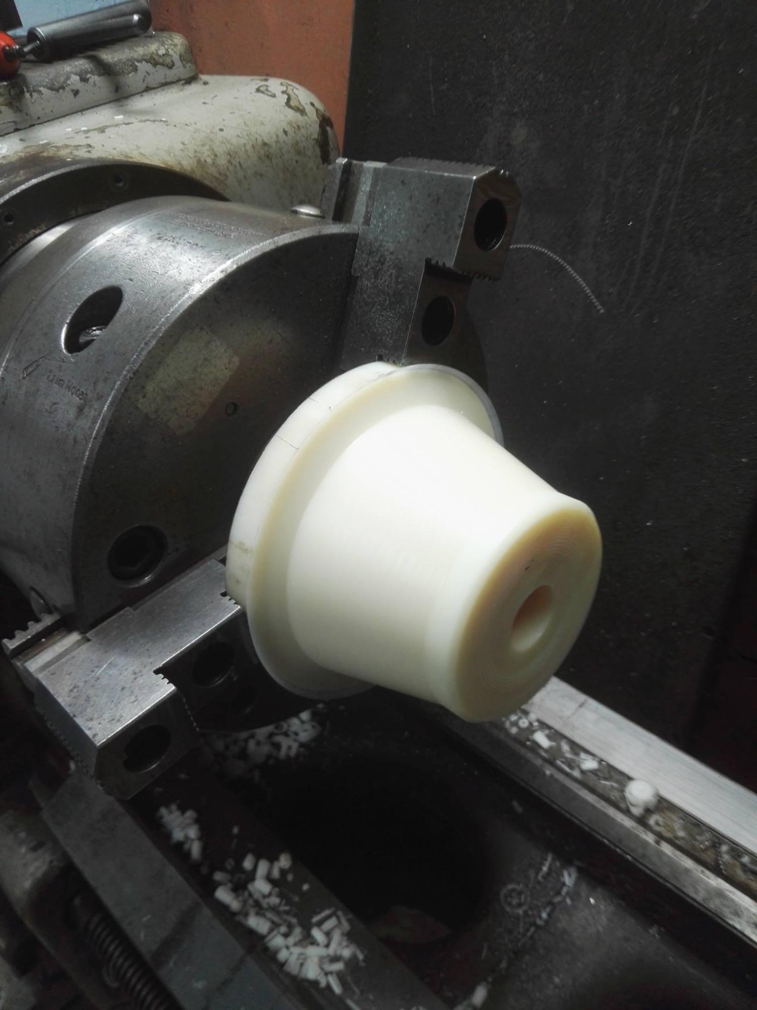 Biały element z tworzywa sztucznego, przypominający stożek, zamocowany w uchwycie tokarki; widoczne wióry i opiłki na prowadnicy tokarki.