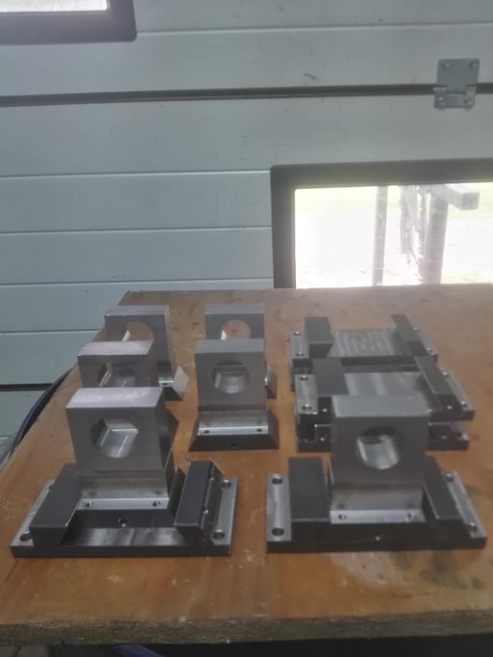 Kilka metalowych uchwytów frezarskich z otworami o różnych kształtach, ułożonych na drewnianym blacie w warsztacie.