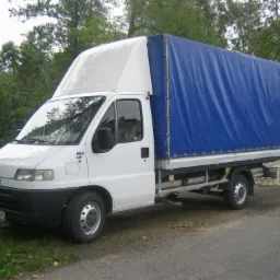 Biały bus dostawczy z niebieską plandeką na tle zieleni i małego budynku. Widoczna tablica rejestracyjna. Ujęcie z boku, lekko z dołu.