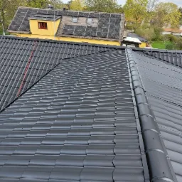 Widok z góry na dwa połączone dachy z czarnej dachówki ceramicznej, jeden z czerwoną rurką wentylacyjną, z żółtym domem w tle z panelami słonecznymi na dachu.