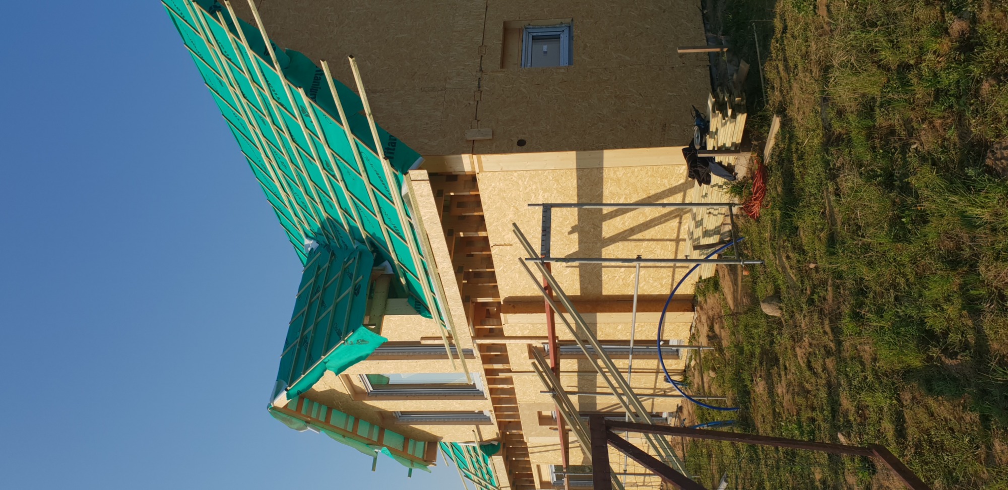 Widok z dołu na szkielet dachu domu jednorodzinnego w trakcie budowy, pokryty zieloną membraną dachową, z widocznymi łatami i kontrłatami, oraz fragmentem elewacji z płyty OSB i rusztowaniem.