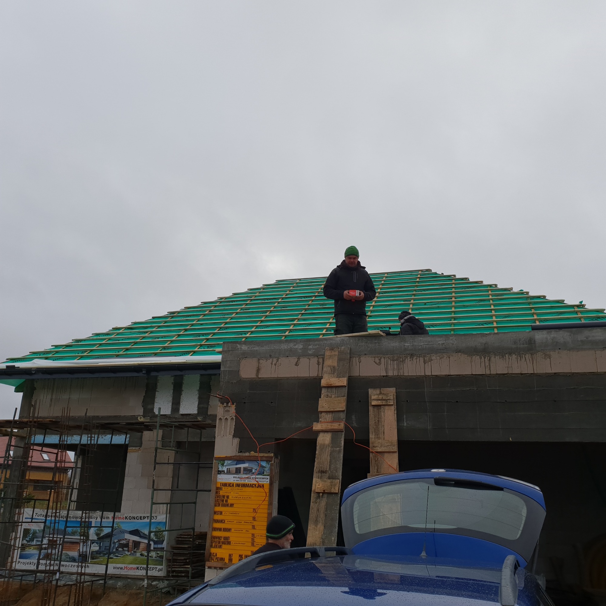 Prace dekarskie na budowie domu jednorodzinnego: widok na zieloną membranę dachową z łatami, dwóch dekarzy na dachu, budynek w stanie surowym otwartym, w tle pochmurne niebo.