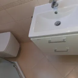 Nowoczesna, biała umywalka z szafką i chromowaną baterią, obok toaleta i fragment kabiny prysznicowej, całość w łazience wykończonej beżowymi płytkami.