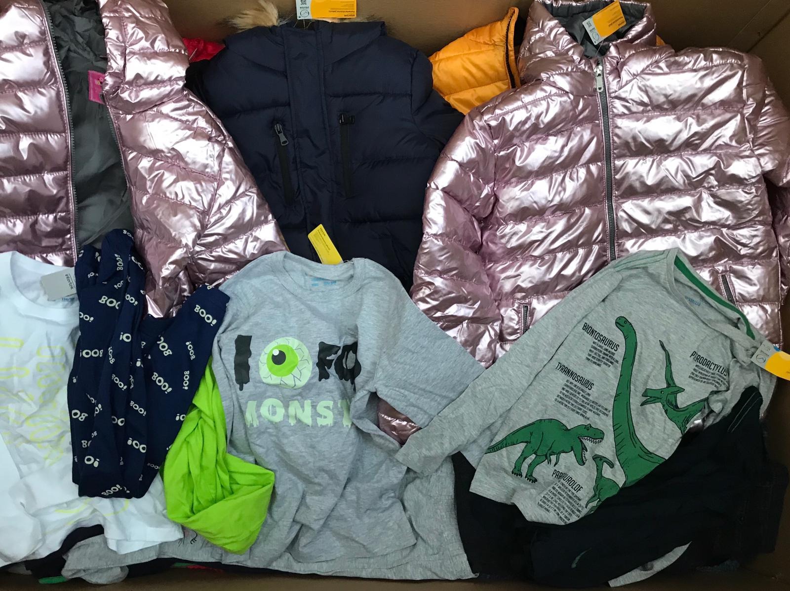 Karton wypełniony różnorodną odzieżą: błyszczące, różowe kurtki puchowe, granatowa kurtka zimowa z futrzanym kapturem, żółta kurtka, koszulki z nadrukami (potwory, dinozaury) oraz ubrania z motywem...