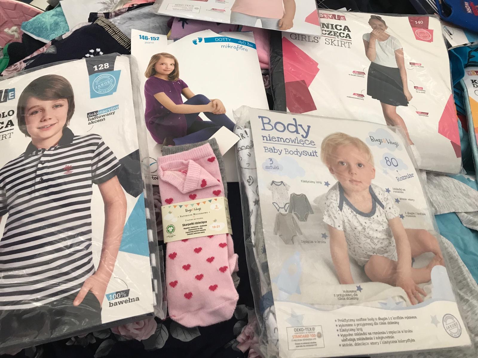 Zestaw zapakowanych ubrań dziecięcych i młodzieżowych, w tym polo w paski, rajstopy, spódniczka, body niemowlęce i skarpetki w serduszka, ułożone na ciemnym tle.