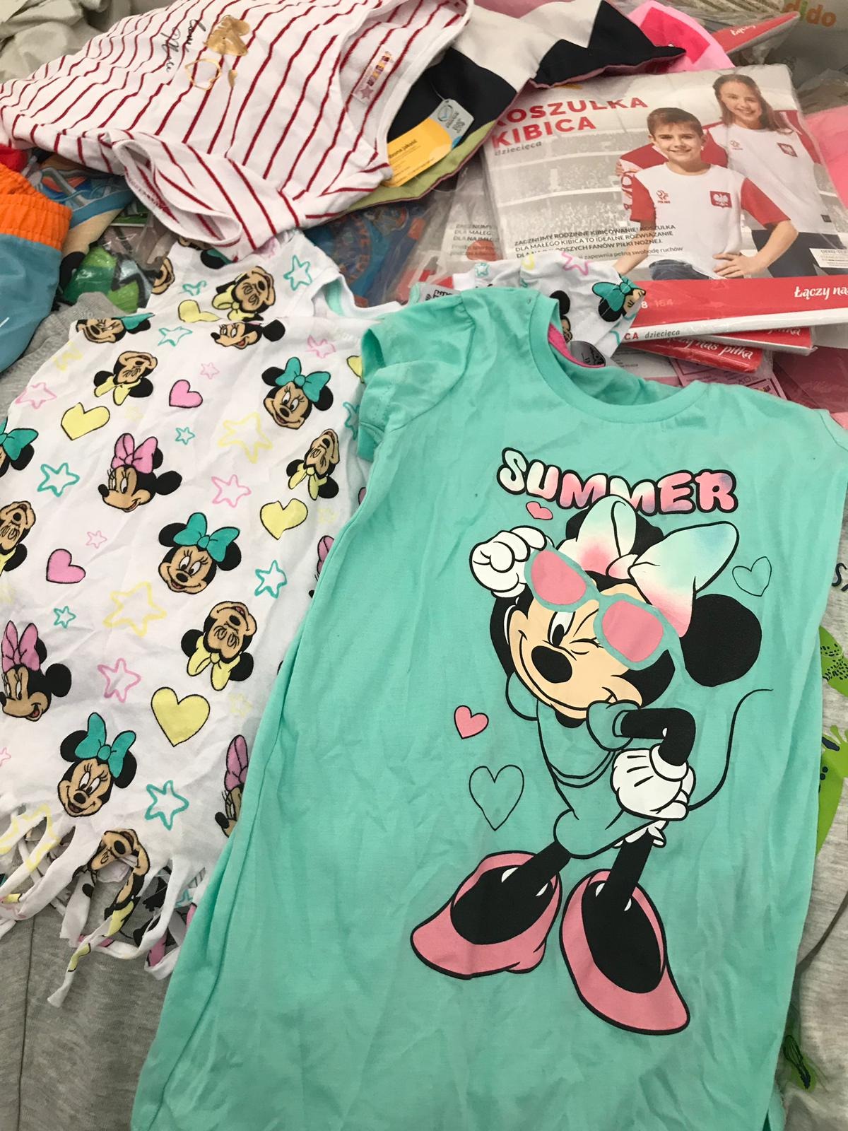 Stos ubrań dziecięcych, w tym turkusowa koszulka z nadrukiem Myszki Minnie w okularach przeciwsłonecznych z napisem 'SUMMER' oraz biała sukienka z motywem Myszki Minnie i elementami frędzli na dole...