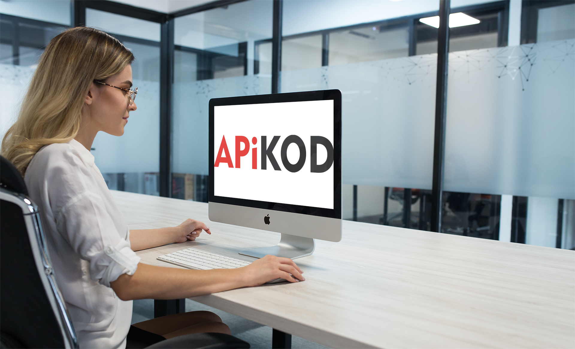 Kobieta w okularach pracuje przy komputerze iMac, na ekranie widoczny napis APIKOD, tło stanowi biuro z przeszklonymi ścianami ozdobionymi geometrycznym wzorem.