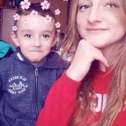 Portret kobiety w czerwonym swetrze i chłopca w szarej bluzie z kapturem, oboje z filtrem kwiatowym na głowach, na tle pokoju z regałem.