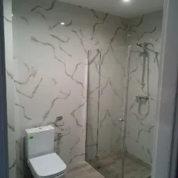 Wykończona łazienka z kabiną prysznicową ze szklanymi drzwiami, toaletą i płytkami imitującymi marmur na ścianach oraz drewnopodobnymi na podłodze.