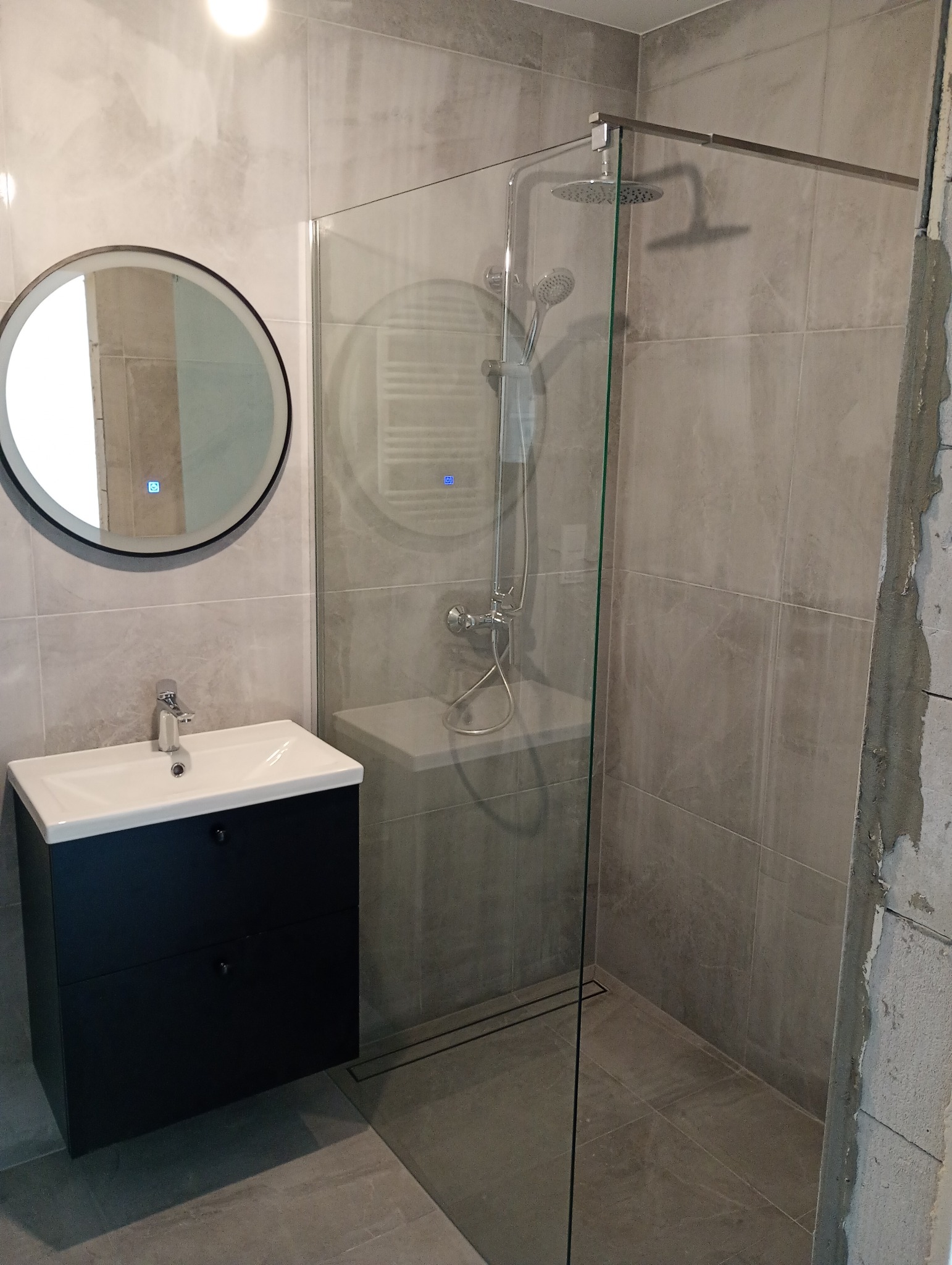 Nowoczesna łazienka z kabiną prysznicową typu walk-in, umywalką z szafką w kolorze czarnym i okrągłym lustrem z podświetleniem LED. Szare płytki imitujące kamień.