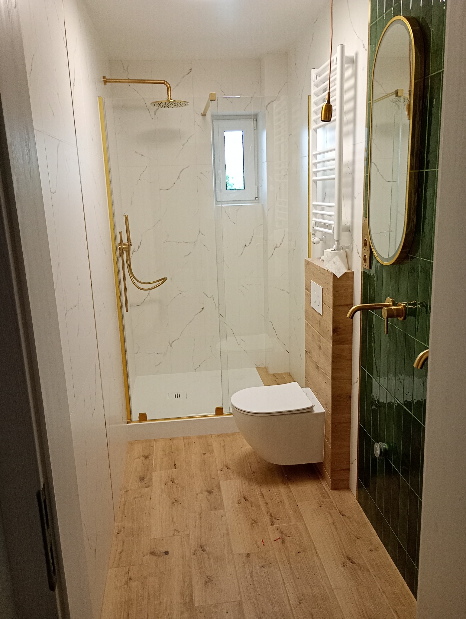 Elegancka łazienka po metamorfozie: kabina walk-in ze złotymi akcentami, toaleta podwieszana, zielone płytki i drewnopodobne panele podłogowe.