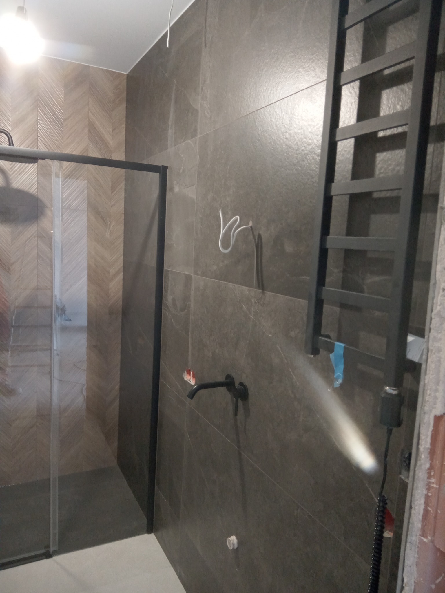 Nowoczesna łazienka z szarą, dużą płytką, czarną armaturą i grzejnikiem drabinkowym. Kabina prysznicowa z czarną ramą. Akcent drewnopodobnych płytek w strefie prysznica.