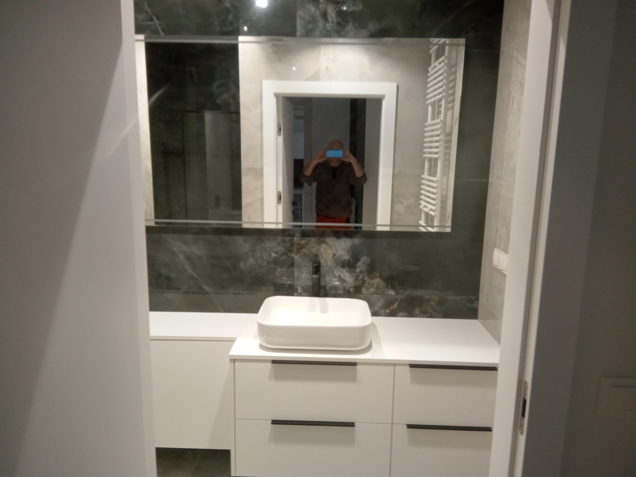 Nowoczesna łazienka po remoncie: biała umywalka nablatowa na szafce z szufladami, lustro z odbiciem osoby robiącej zdjęcie, ciemne płytki na ścianie.